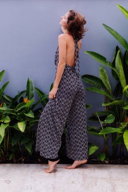 Arkadaki duvar ve tropikal yapraklarda poz veren klasik uzun tulumlu Kafkas tarzı model kadının moda portresi.