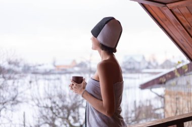 Kafasında havluyla, elinde çay tutan genç bir kadın kışın balkonda duruyor.