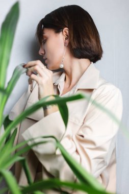 Beyaz tenli, bej takım elbiseli, sütyensiz, palmiye ağacının arkasında gri arka planda poz veren yumuşak bir kadın portresi..