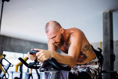 Dövmeli güçlü, kaslı, sakallı Avrupalı adam spor salonunda spor yapıyor. Büyük pencerenin yanında, dışarıda ağaçların manzarası var.