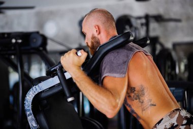 Avrupa dövmeli, sakallı, güçlü kaslı bir adam eğitim makinesine çömeliyor, spor aletleri..