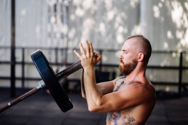 Spor salonunda sakallı, güçlü bir adam dövmesi var. Halter egzersizi yapan kaslı vücut geliştirici.
