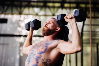 Genç, kaslı, güçlü, kaslı, sakallı Avrupalı adam omuzları için halterlerle sıkı egzersiz yapıyor.