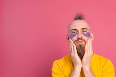 Sarı tişörtlü ve mor göz bandı maskeli Avrupalı yakışıklı adam, erkekler için yüz bakımı yapıyor. Pembe arka planda izole edilmiş.