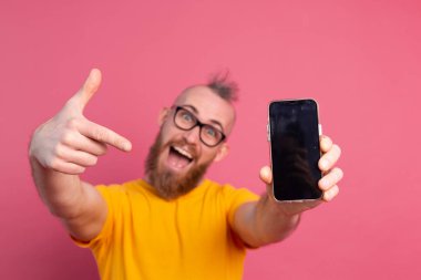 Hey yeni bir şey. Cep telefonunu siyah ekranla gösteren mutlu Avrupalı sakallı adam. Pembe arka plan izole edilmiş, boş alan.