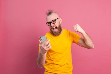 Çığlık atan genç sakallı yakışıklı bir adam resmi. Pembe duvarın arkasında izole bir şekilde duruyor. Cep telefonu kullanarak kazanan hareketi yap.