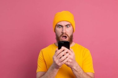 Portrait of concerned shocked mature bearded male gasping unhappy holding smartphone reading strange and disturbing message, isolated pink wall