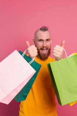 Mutlu sakallı, sıradan görünüşlü, renkli alışveriş çantaları olan, stüdyo pembe arka planlı, mutlu bir adam.