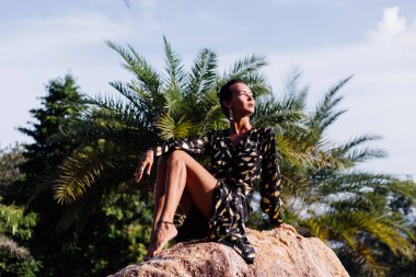 Fashion portrait of slim tanned fit short dark hair woman in black golden long cape on tropical road near palm trees