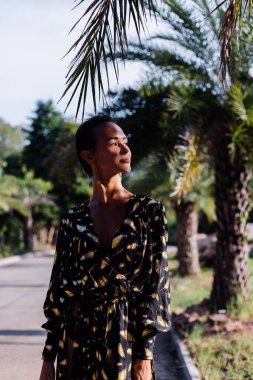 Fashion portrait of slim tanned fit short dark hair woman in black golden long cape on tropical road near palm trees
