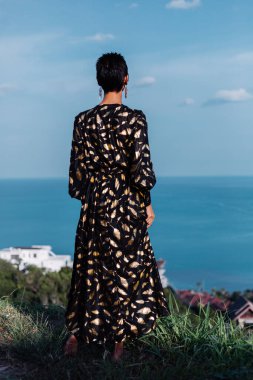 Portrait of beautiful luxury look fit tanned slim woman in black golden cape and sunglasses on top of mountain with amazing sea view in Thailand