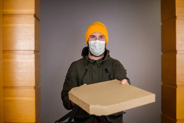 Caucasian deliver man wearing face medical mask handling paper box with pizza inside, give to costumer in doorway