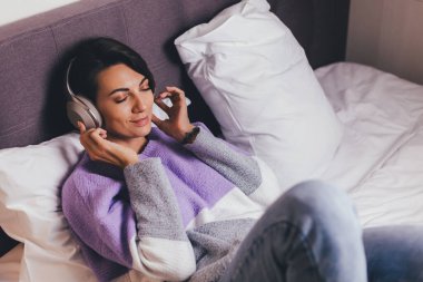 Mutlu kadın rahat yatağında sıcak kıyafetler giyiyor, kazak giyiyor, müzik dinliyor.