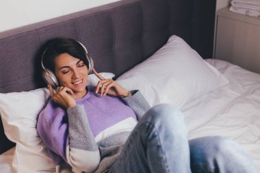 Mutlu kadın rahat yatağında sıcak kıyafetler giyiyor, kazak giyiyor, müzik dinliyor.