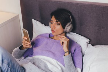 Mutlu kadın rahat yatağında sıcak kıyafetler giyiyor, kazak giyiyor, müzik dinliyor.