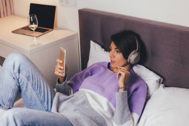 Mutlu kadın rahat yatağında sıcak kıyafetler giyiyor, kazak giyiyor, müzik dinliyor.