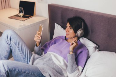 Mutlu kadın rahat yatağında sıcak kıyafetler giyiyor, kazak giyiyor, müzik dinliyor.