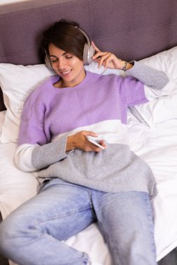 Mutlu kadın rahat yatağında sıcak kıyafetler giyiyor, kazak giyiyor, müzik dinliyor.