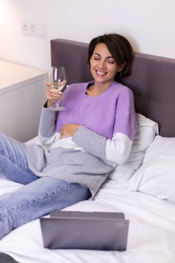 Happy woman in warm pullover in bed with glass of wine alone by herself resting watching movie comedy smile laug