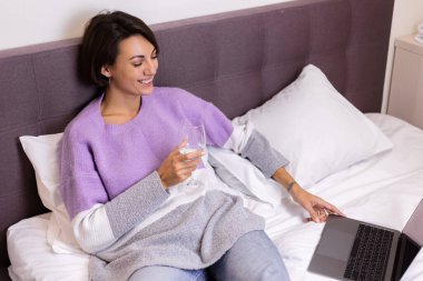 Happy woman in warm pullover in bed with glass of wine alone by herself resting watching movie comedy smile laug