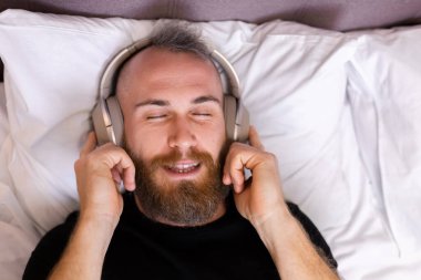 Happy caucasian man on bed wearing headphone listen enjoy his favorite music, resting alone, dancing