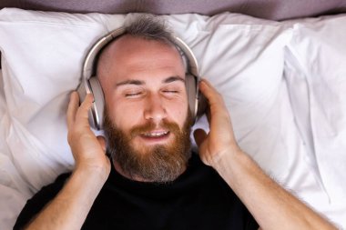 Happy caucasian man on bed wearing headphone listen enjoy his favorite music, resting alone, dancing
