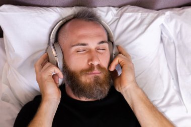 Happy caucasian man on bed wearing headphone listen enjoy his favorite music, resting alone, dancing