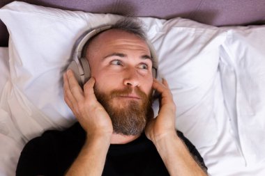 Happy caucasian man on bed wearing headphone listen enjoy his favorite music, resting alone, dancing