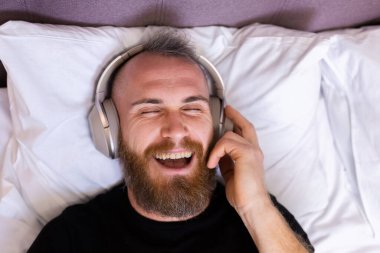 Happy caucasian man on bed wearing headphone listen enjoy his favorite music, resting alone, dancing
