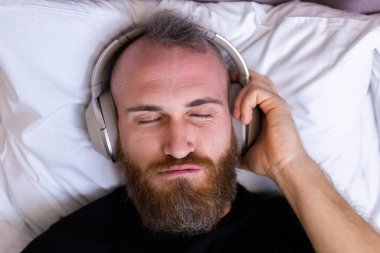 Happy caucasian man on bed wearing headphone listen enjoy his favorite music, resting alone, dancing