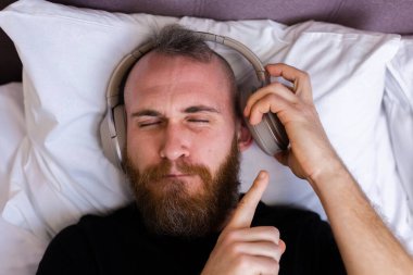 Happy caucasian man on bed wearing headphone listen enjoy his favorite music, resting alone, dancing