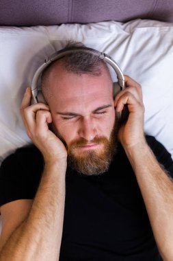 Happy caucasian man on bed wearing headphone listen enjoy his favorite music, resting alone, dancing