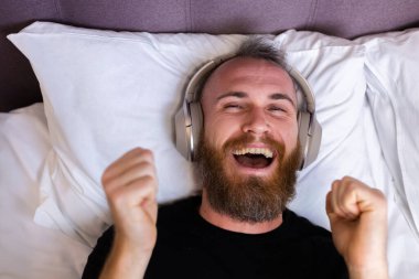 Happy caucasian man on bed wearing headphone listen enjoy his favorite music, resting alone, dancing