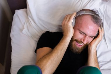 Happy caucasian man on bed wearing headphone listen enjoy his favorite music, resting alone, dancing