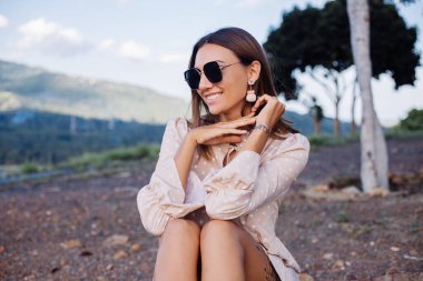Romantic outdoor close portrait of young calm positive caucasian tanned woman with sunglasses and earings at warm sunset in park