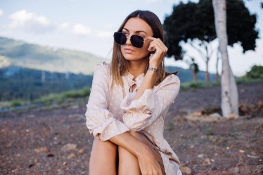 Romantic outdoor close portrait of young calm positive caucasian tanned woman with sunglasses and earings at warm sunset in park
