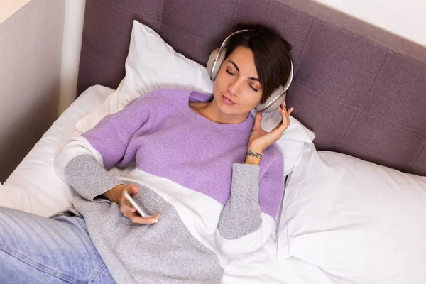 Mutlu kadın rahat yatağında sıcak kıyafetler giyiyor, kazak giyiyor, müzik dinliyor.