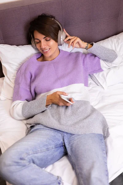 Mutlu kadın rahat yatağında sıcak kıyafetler giyiyor, kazak giyiyor, müzik dinliyor.