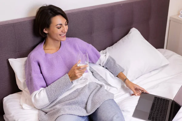 Happy woman in warm pullover in bed with glass of wine alone by herself resting watching movie comedy smile laug