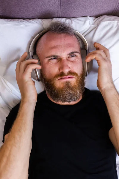 Happy caucasian man on bed wearing headphone listen enjoy his favorite music, resting alone, dancing