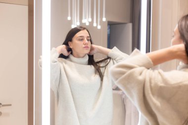 Young beautiful woman in a sweater in a beauty salon looks in the mirror, touches her face, thinks about the upcoming procedures, considers herself