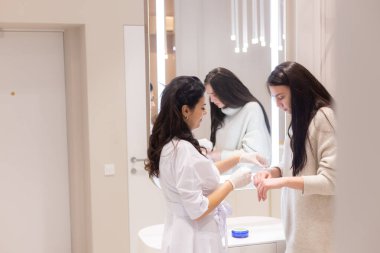 Two women, a beautician doctor and a client, stand at the mirror, for a consultation, the doctor applies cream to the woman's hands and makes a light massage