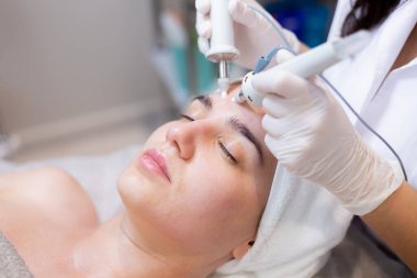 Genç ve güzel bir kız güzellik uzmanının masasına uzanır ve cilt gençleştirme ve nemlendirme için profesyonel bir aletle işlem görür.