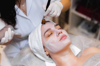 Fırçalı bir güzellik uzmanı güzellik salonundaki genç bir kız müşterinin yüzüne beyaz nemlendirici bir maske takıyor.