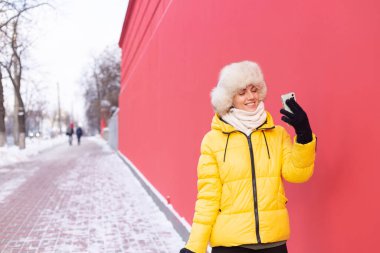 Kış güneşli bir günde, kırmızı bir duvarın arkasında mutlu bir genç kadın. Karlı bir kaldırımda gülümsüyor ve telefonda konuşuyor.