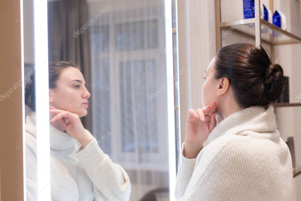 Young beautiful woman in a sweater in a beauty salon looks in the mirror, touches her face, thinks about the upcoming procedures, considers herself