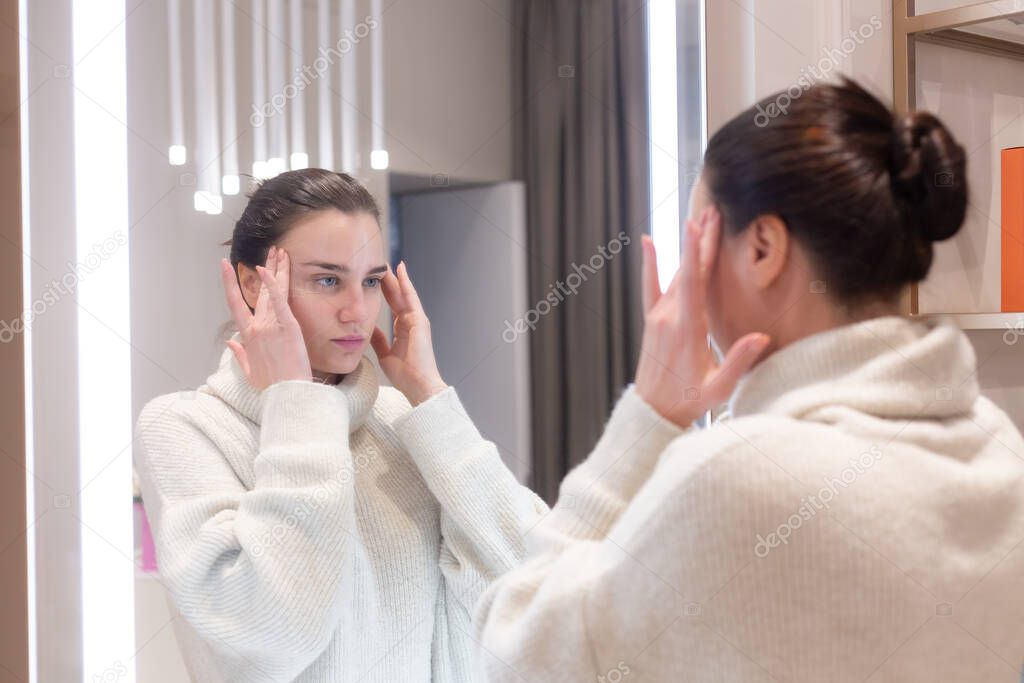 Young beautiful woman in a sweater in a beauty salon looks in the mirror, touches her face, thinks about the upcoming procedures, considers herself