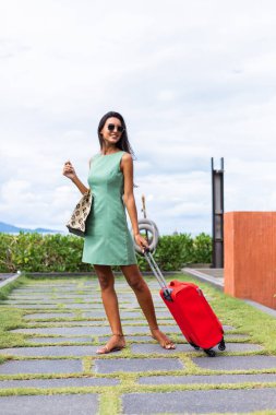 Happy caucasian pretty long hair elegant tourist woman in dress with red suitcase outside hotel on vacation