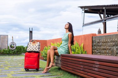 Happy caucasian pretty long hair elegant tourist woman in dress with red suitcase outside hotel on vacation