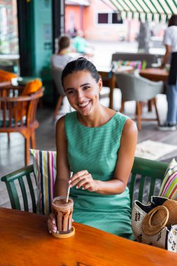 Cute elegant calm happy woman in green summer dress sits with coffee in cafe enjoying morning